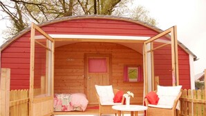 A wooden cabin with a red exterior, a porch with wicker chairs and a small table, and a bed with a floral bedspread. - Bleckmanns Hof (Werne)