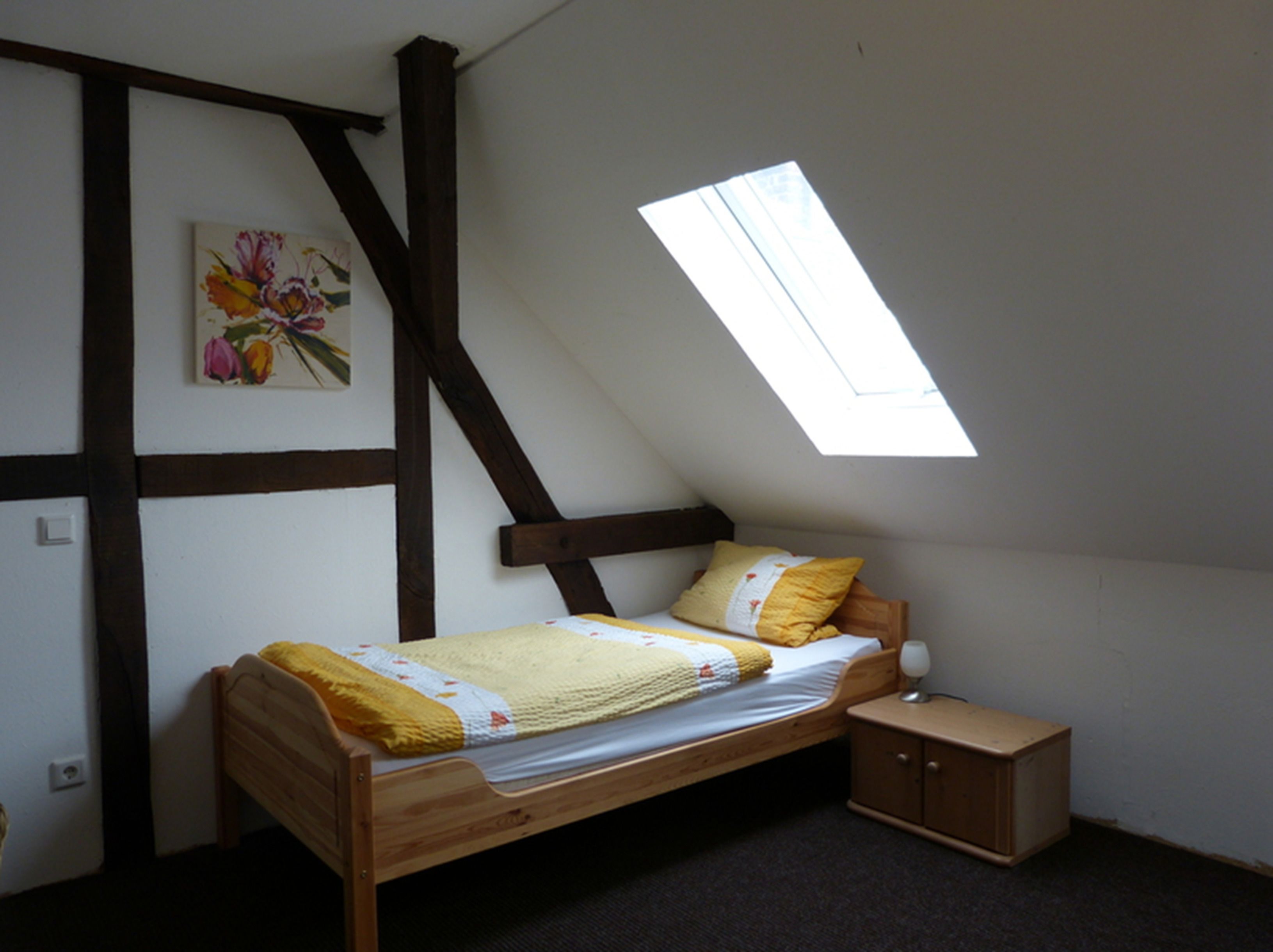 A small, cozy bedroom with a sloped ceiling and a skylight. It features a single bed with a yellow blanket and a small wooden nightstand.