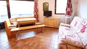 A cozy living room with a wooden bench, a sofa, a TV on a wooden cabinet, and large windows with curtains. - Spacious 3-Room Apartment for 8, 400m from Lift, Parking, Ski Locker, Near Amenities (La Salle-les-Alpes)