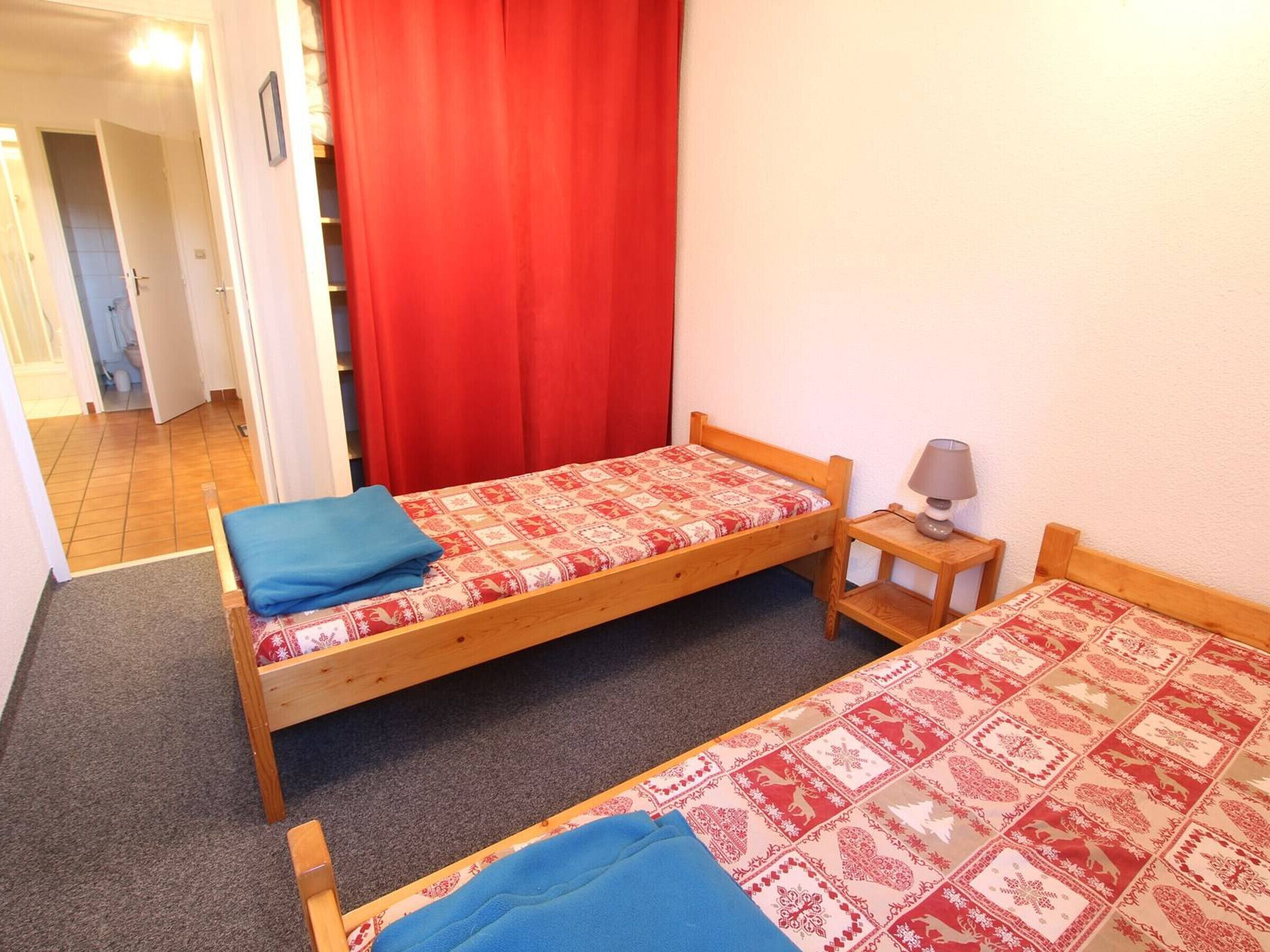A small, simple room with two wooden beds, a red curtain, a small table with a lamp, and a door leading to another room.