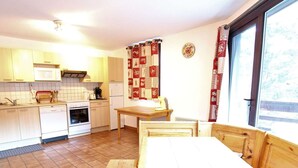 A compact kitchen with a dining area, white cabinets, a wooden table, and a window with curtains. - Spacious 3-Room Apartment for 8, 400m from Lift, Parking, Ski Locker, Near Amenities (La Salle-les-Alpes)
