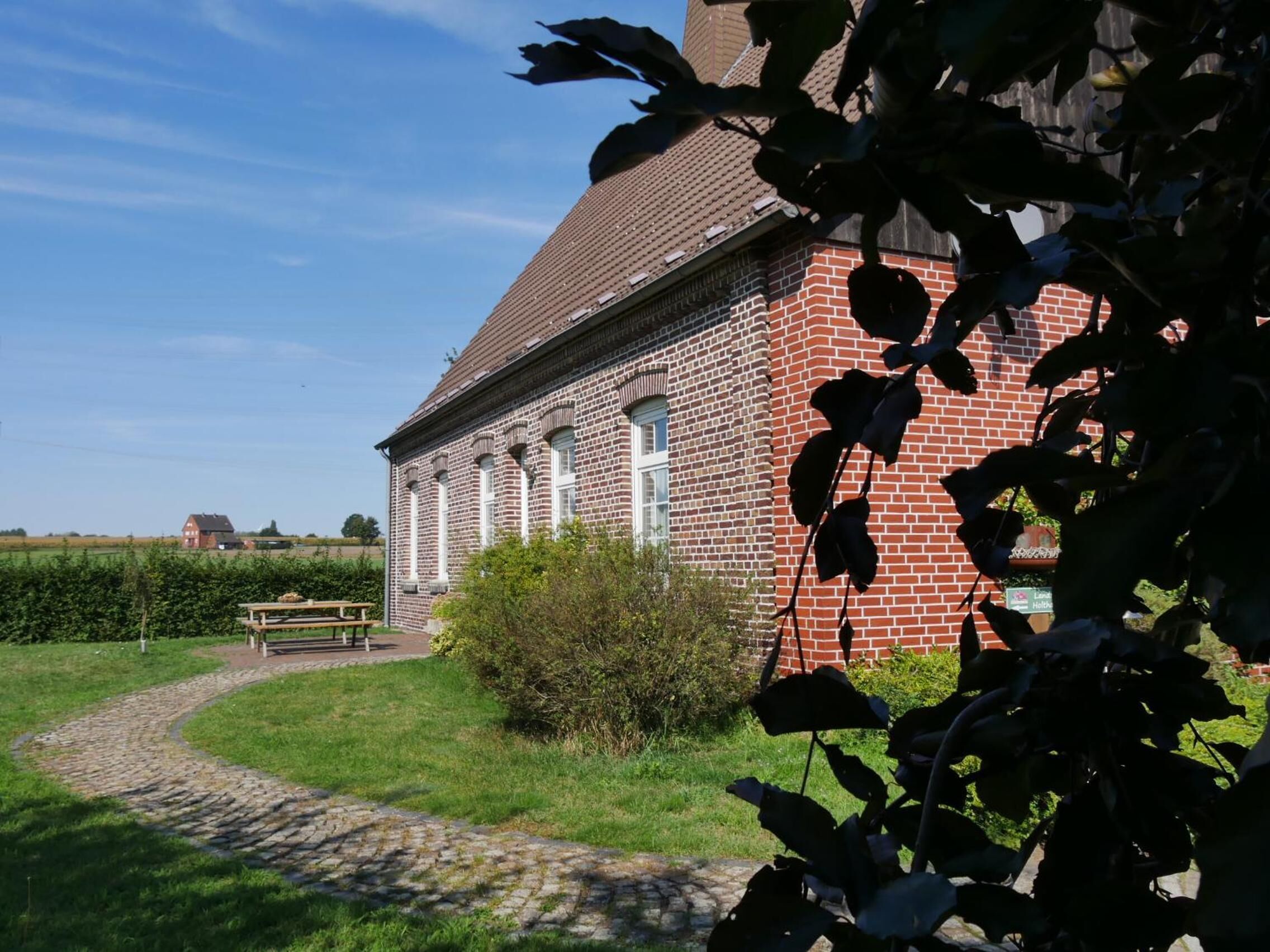A brick building with a sloped roof and white window frames, surrounded by a grassy area and a cobblestone path.