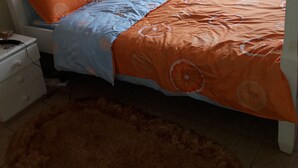 A bed with orange and blue bedding, a fluffy brown rug, and a white bedside table with a lamp. - A home away from home (Ruaka)