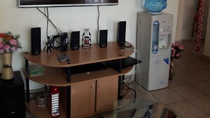 A living room with a flat-screen TV mounted on the wall, a wooden entertainment center with speakers, a water dispenser, and a decorative clock. - A home away from home (Ruaka)