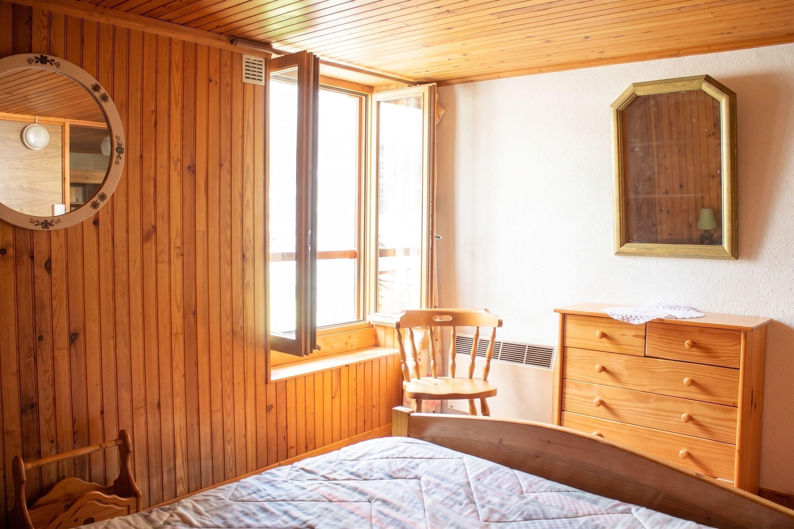 A wooden-paneled room with a bed, a chair, a dresser, a mirror, and a window.