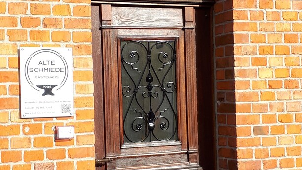 Edificio de ladrillo con puerta de madera y reja metálica. Un letrero en la pared dice "ALTE SCHMIEDE GASTEAUS 1904".