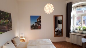 Un dormitorio con una cama, una mesita de noche con lámpara, un espejo en la pared y una ventana con vistas a una casa y jardín.