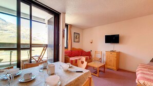 A room with a dining table set for four, a bed with a red patterned bedspread, a television mounted on the wall, and a window with a view of a mountainous landscape. - Spacious apartment for 6, steps from slopes, balcony with Masse view (Les Menuires)