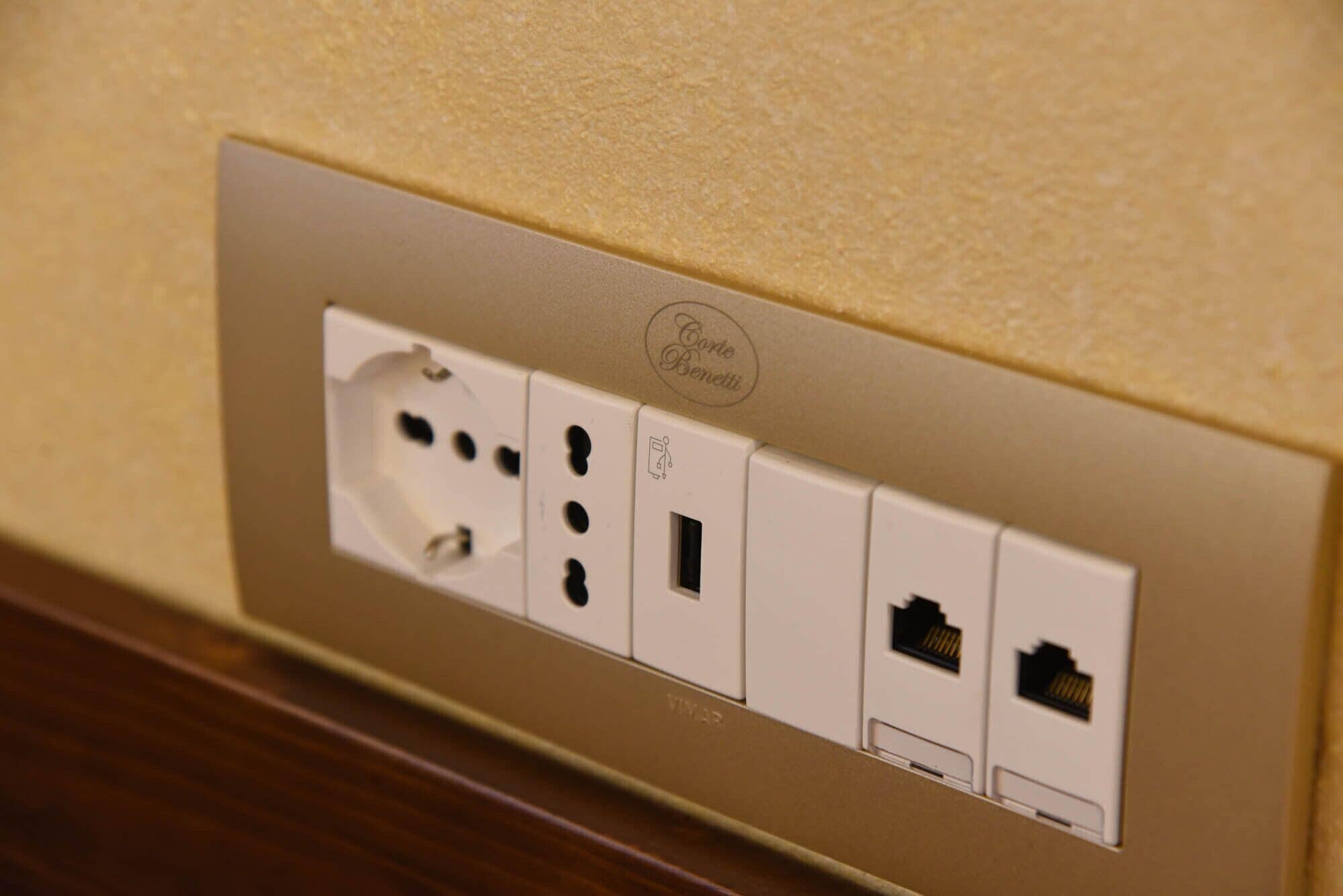 Toma corrientes empotrada en la pared con varios enchufes, incluyendo puertos USB, y una etiqueta que dice 'Corte Benetti'.