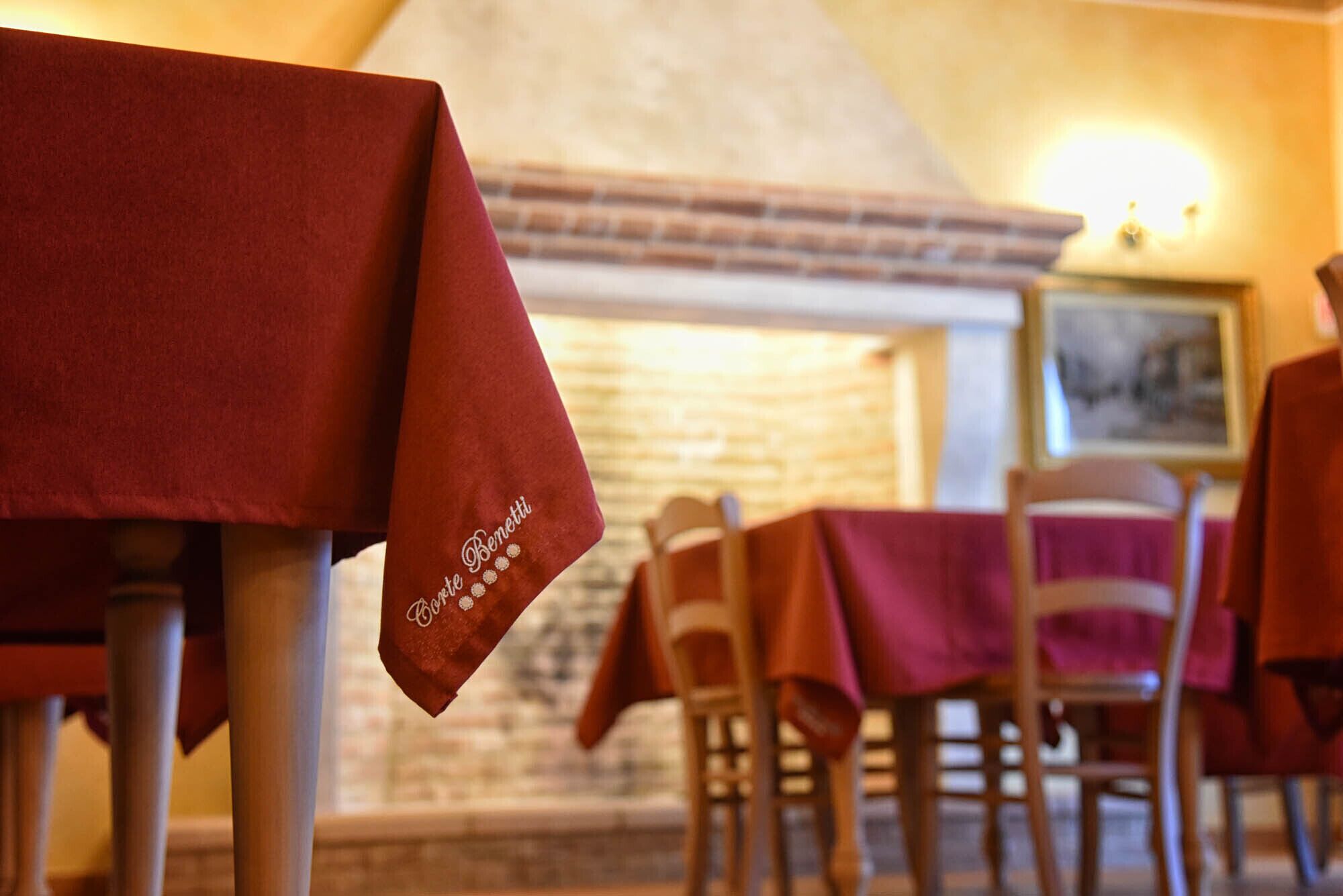 A restaurant interior with tables covered by red tablecloths, wooden chairs, and a brick wall with a fireplace.