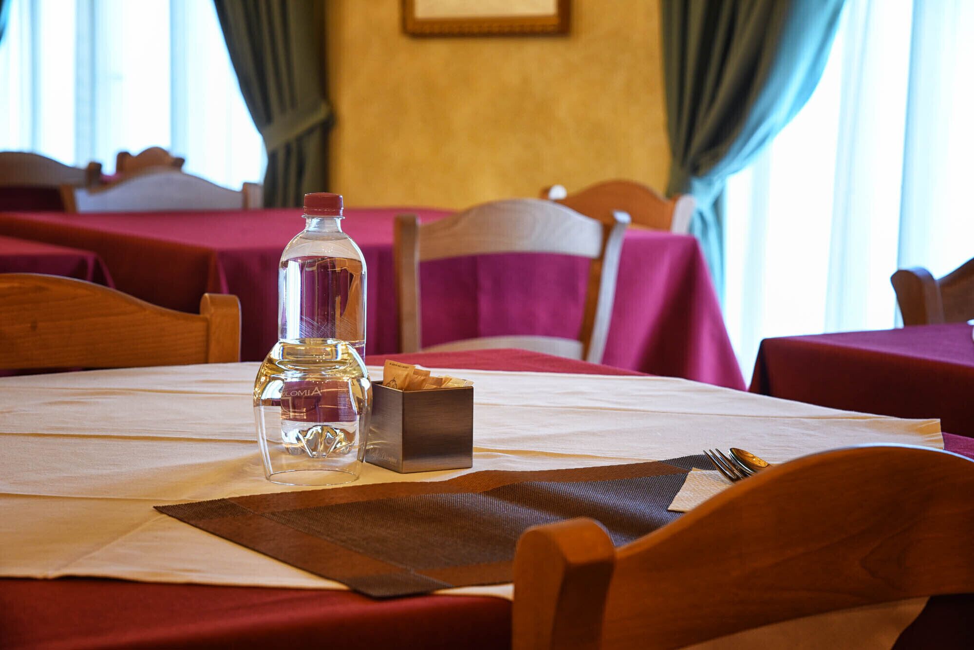 A hotel dining area with tables set for two, featuring wooden chairs and a bottle of water on the table.