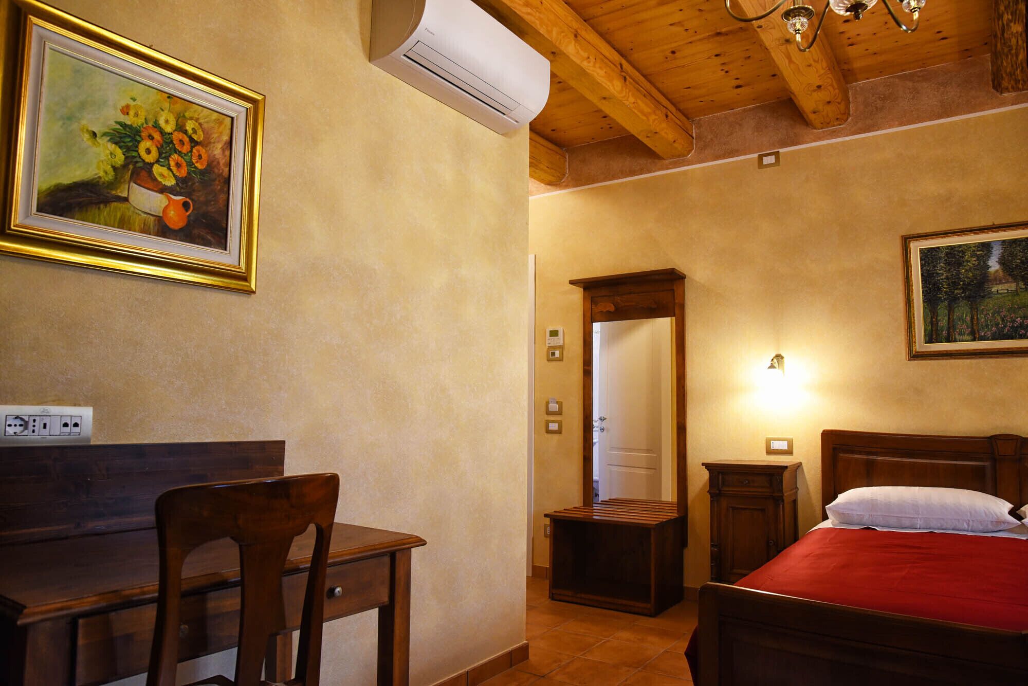A bedroom with a wooden bed, a desk with a chair, a painting on the wall, and an air conditioning unit.