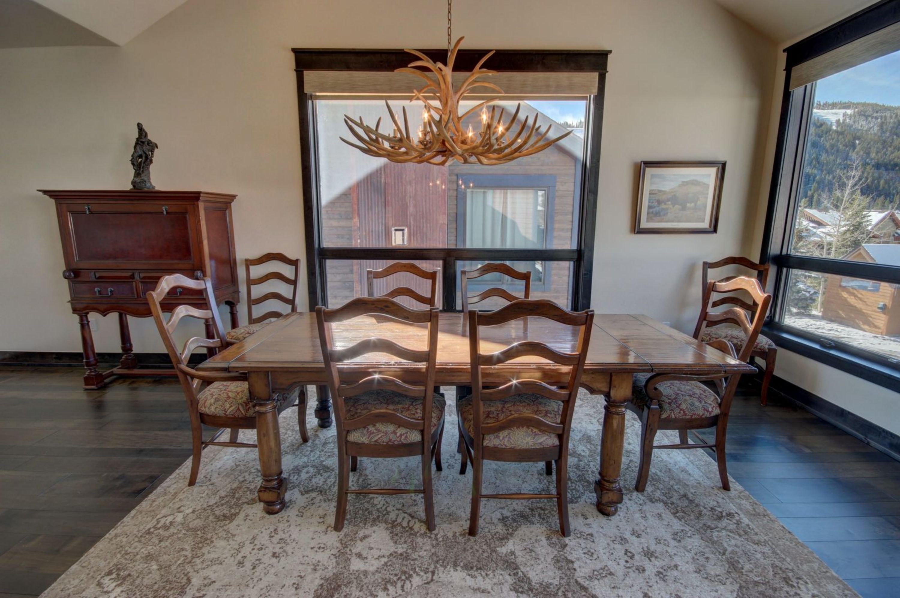 A dining room with a large wooden table, six chairs, a chandelier, and a view of a red barn through a large window.