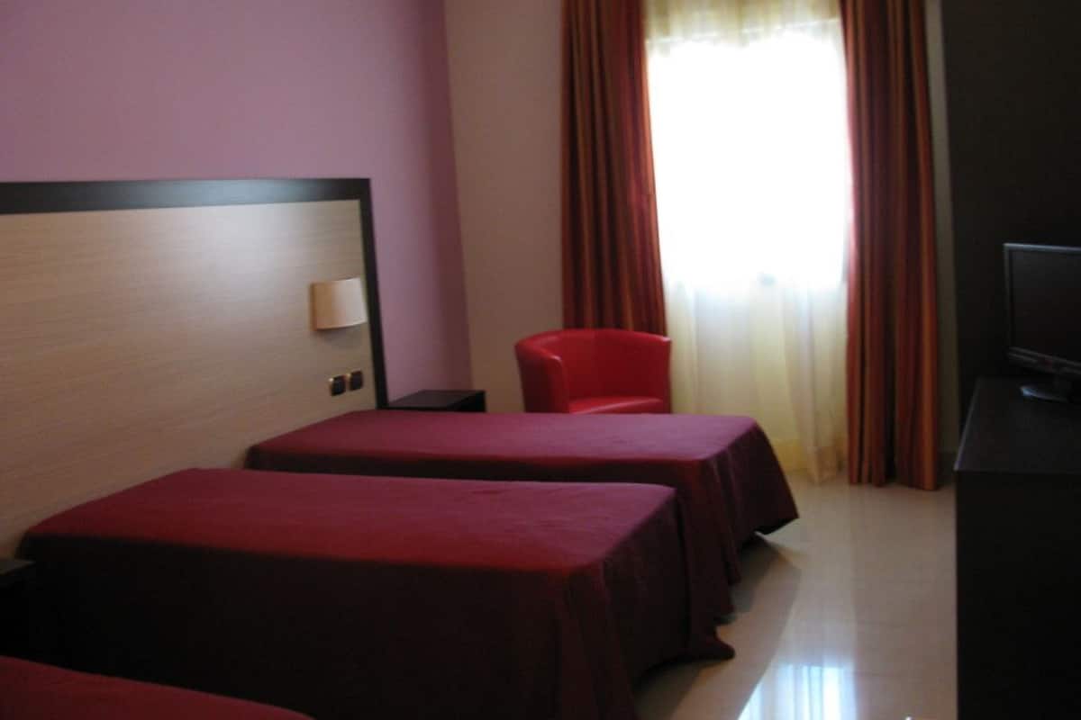 Una habitación de hotel con dos camas, una silla roja, un televisor y una ventana con cortinas.