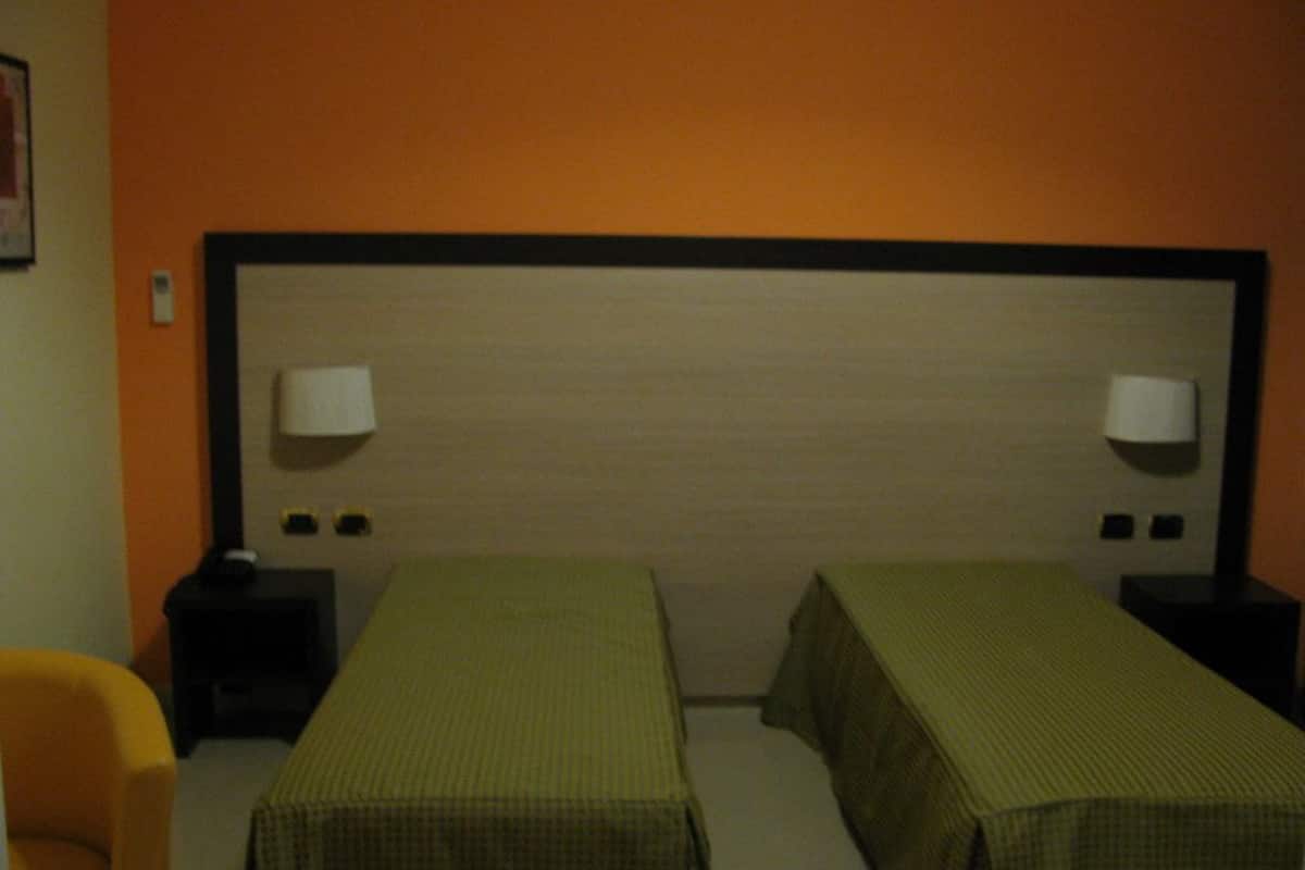 Habitación de hotel con dos camas individuales, una mesita de noche con lámpara y una silla.