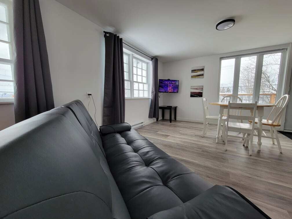 A modern living room with a black leather sofa, a dining table with white chairs, and a TV mounted on the wall.