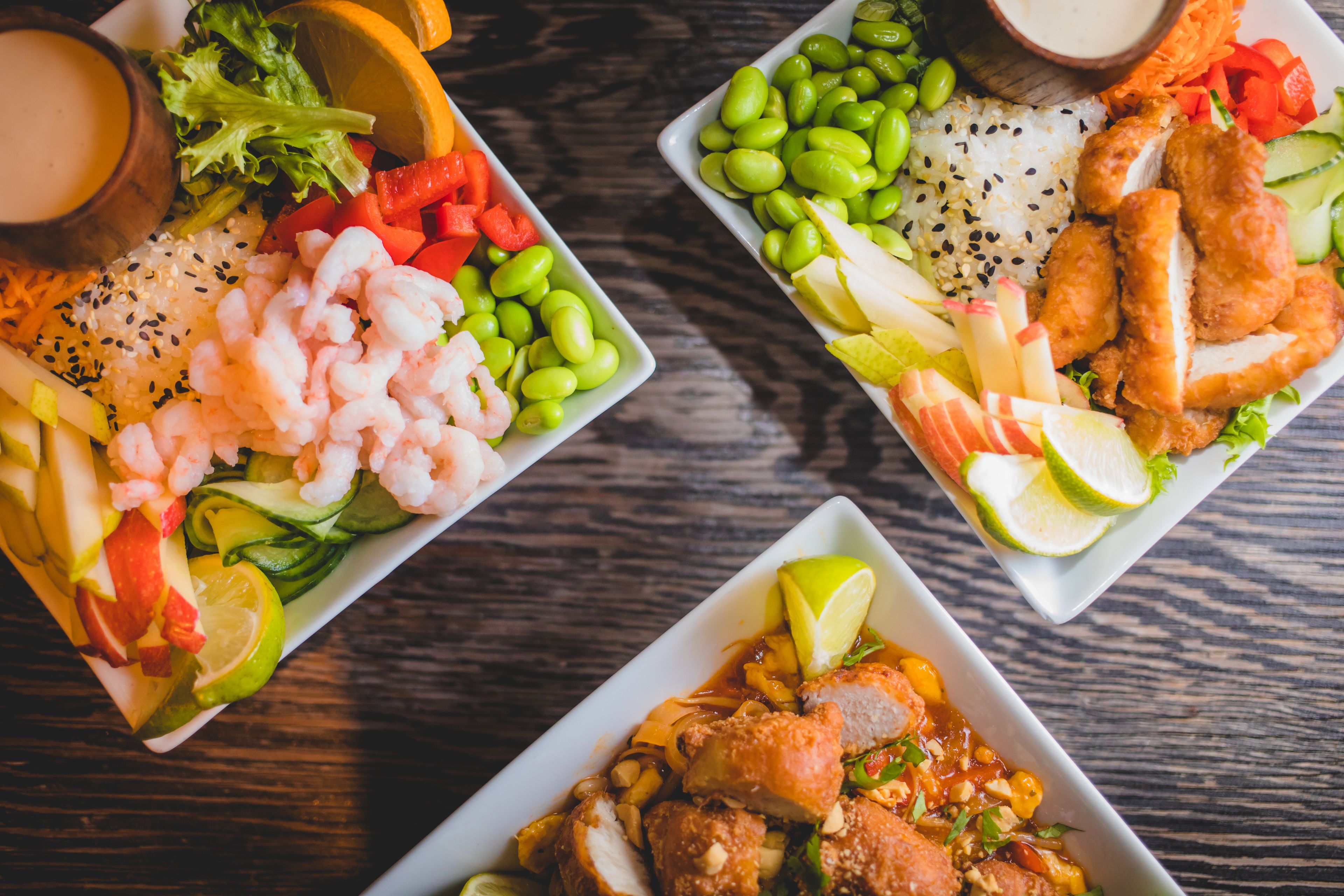 Tres platos cuadrados con diferentes alimentos, como camarones, arroz y verduras, sobre una mesa de madera.