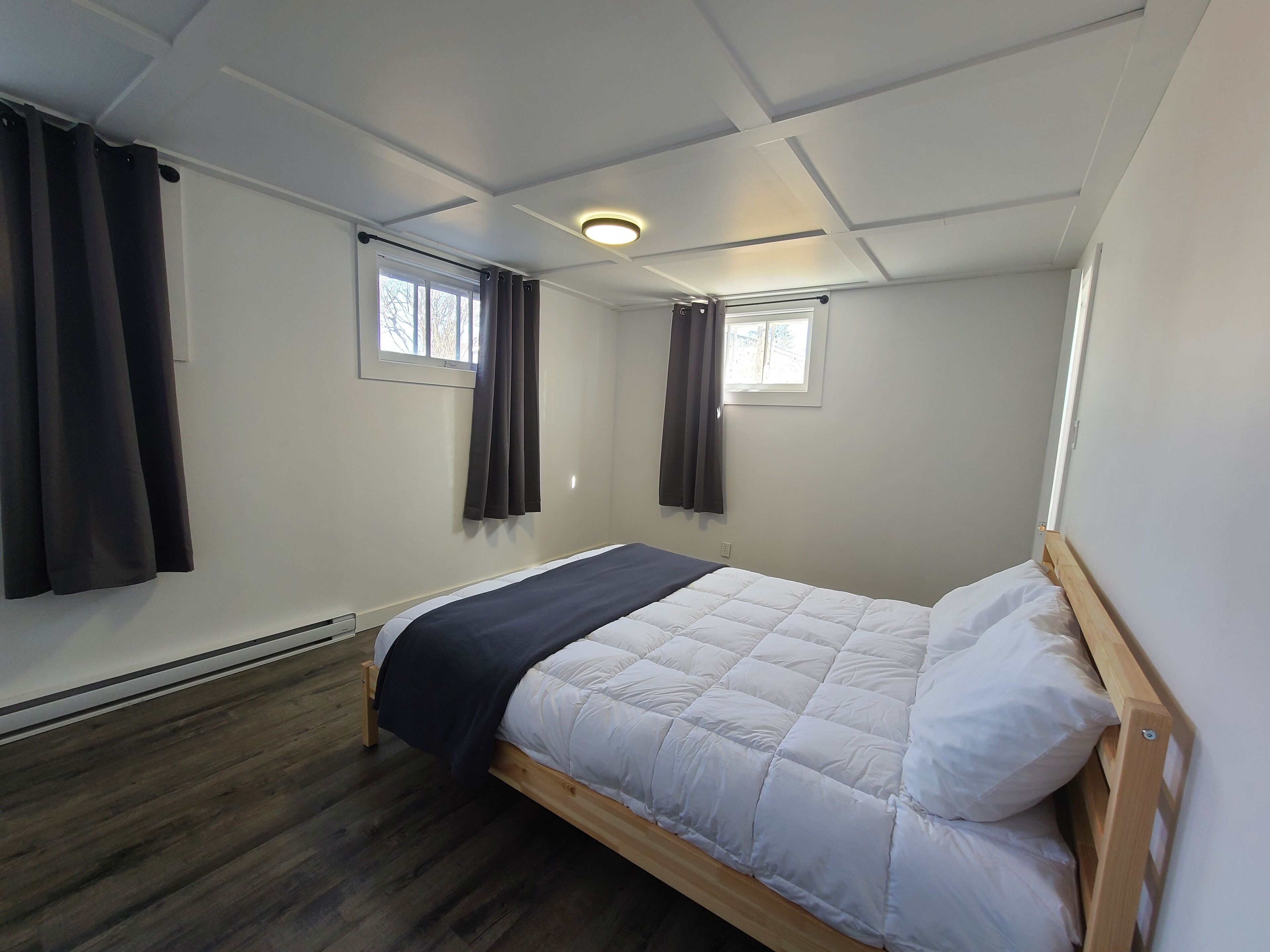Une petite chambre avec un lit simple, un cadre en bois, un matelas matelassé blanc et une couverture de couleur foncée. Il y a deux fenêtres avec des rideaux, un luminaire au plafond et un radiateur au sol.
