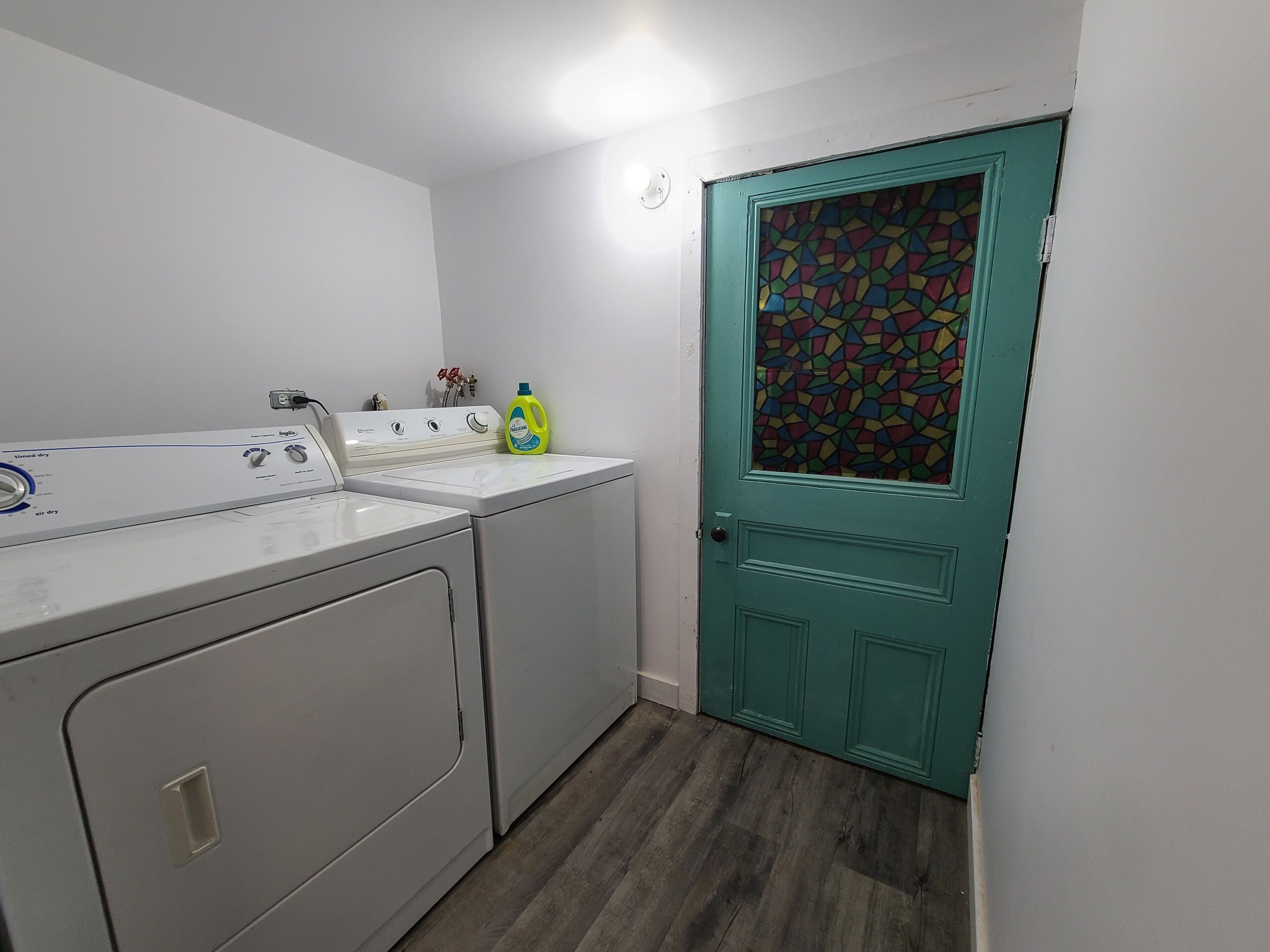 A laundry room with a front-loading washer and dryer, a teal door with a stained glass window, and a bottle of detergent on the dryer.