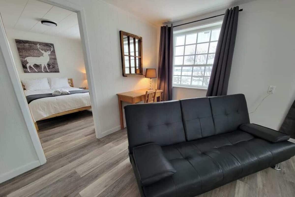 Ett modernt rum med en svart lädersoffa, ett skrivbord i trä, en säng med ett vitt sänggavel och ett stort fönster med gardiner.