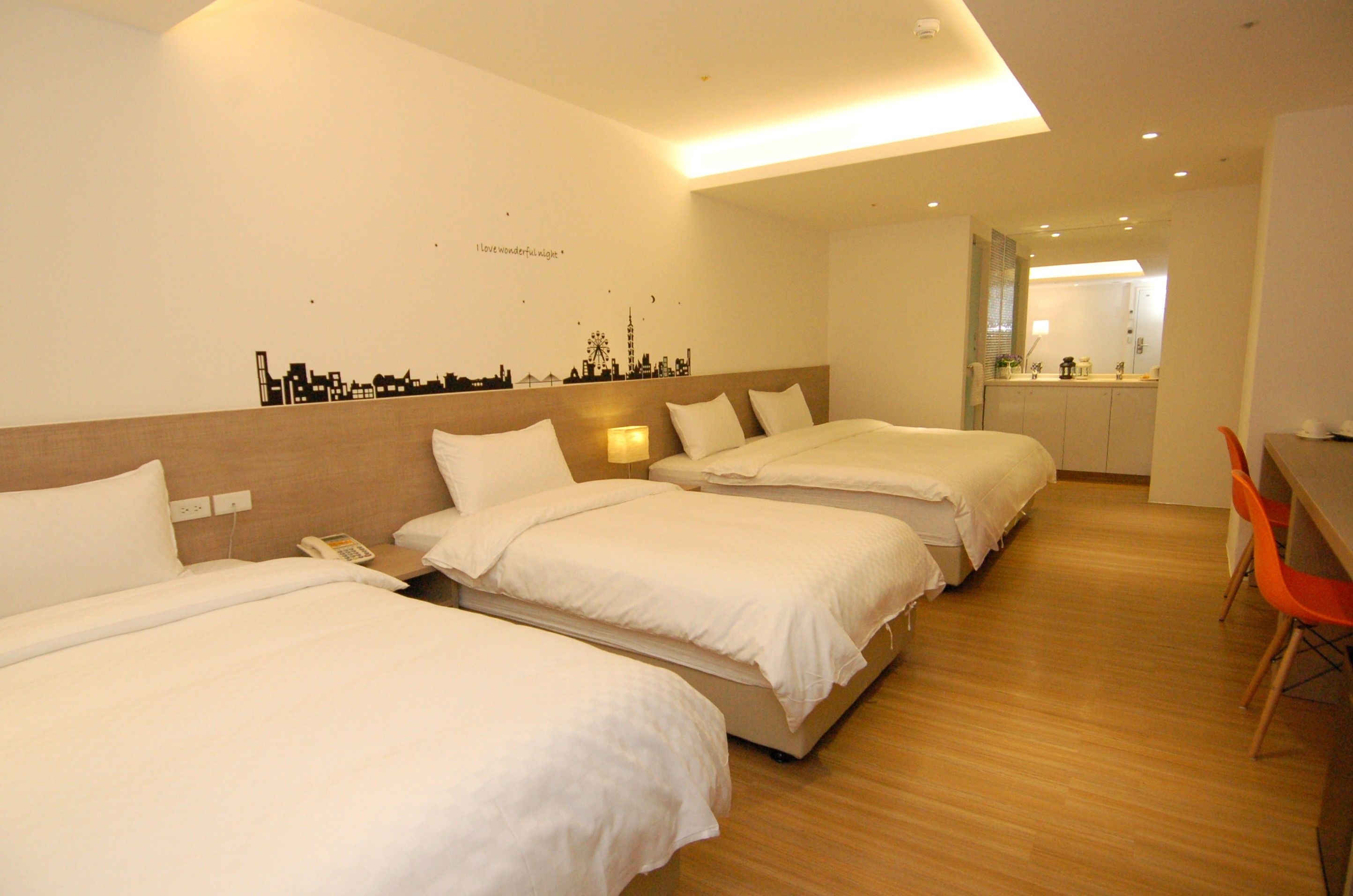 Habitación de hotel con tres camas, un escritorio y una silla. Se observa un cuadro con un paisaje urbano y un espejo en la habitación.