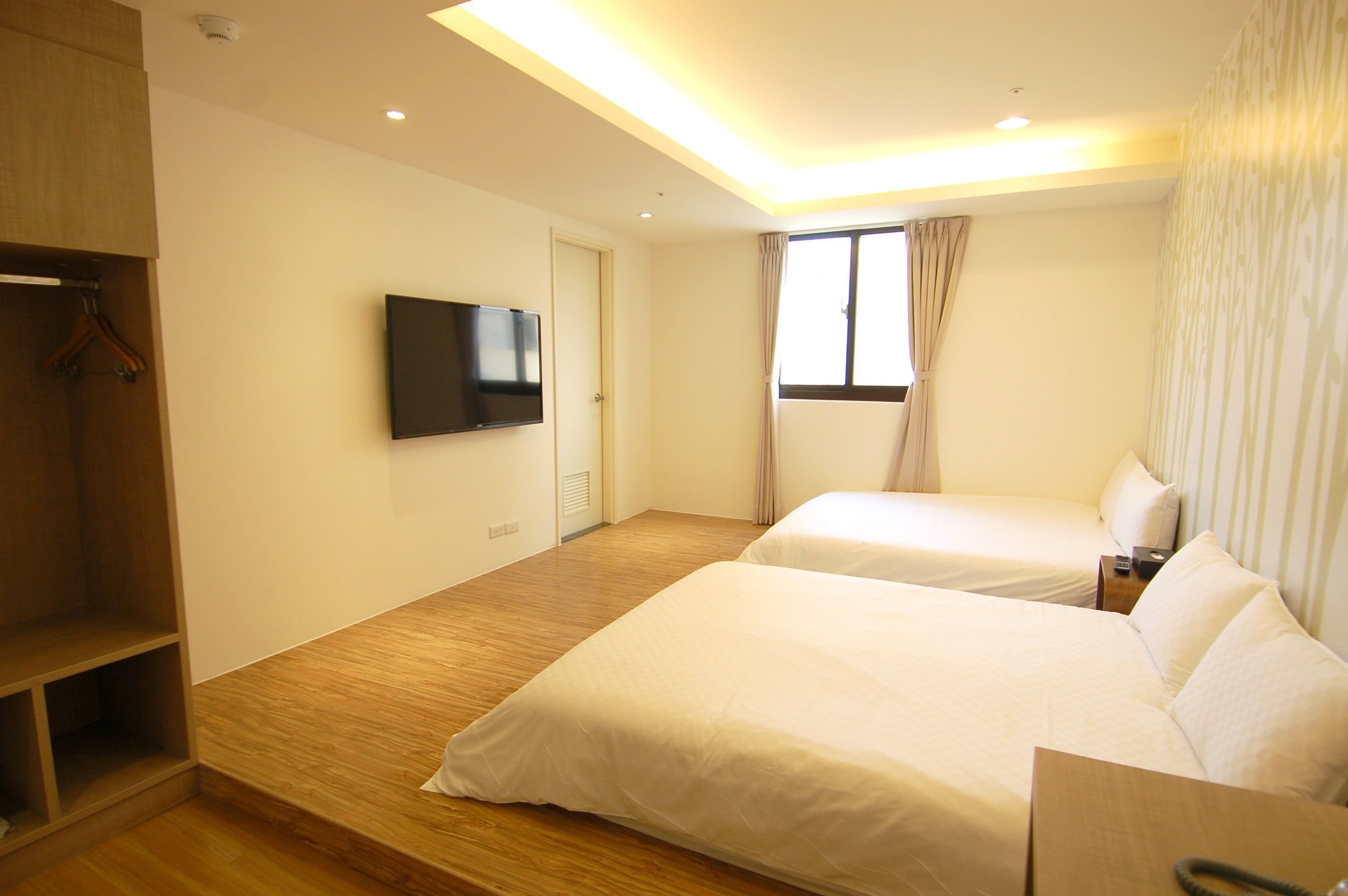 A hotel room with two beds, a TV mounted on the wall, a wooden floor, and a window with curtains.