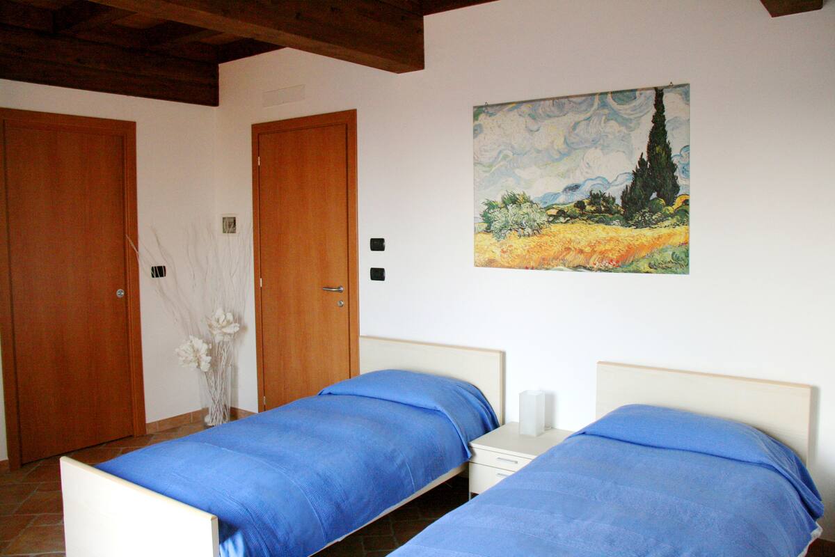 Un dormitorio con dos camas individuales, puertas de madera, un cuadro en la pared y un florero con flores blancas.