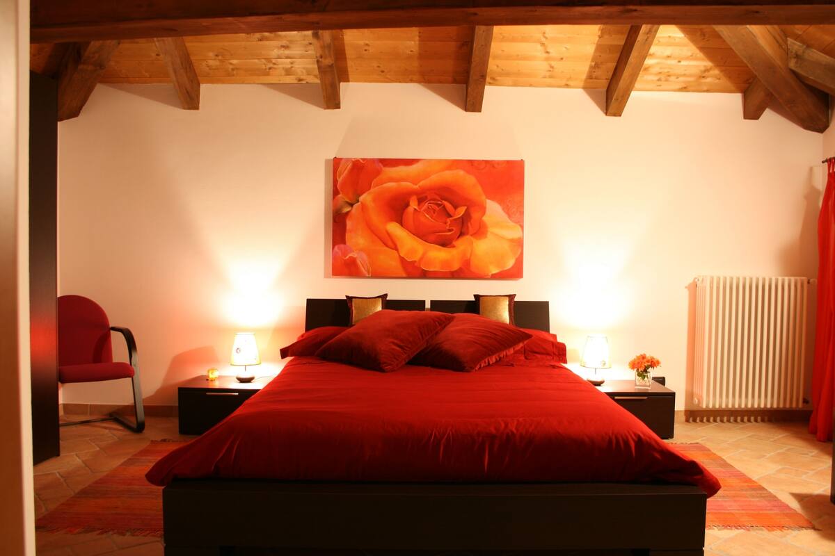 Un dormitorio con techo de madera, una cama grande con ropa de cama roja, dos mesitas de noche con lámparas y una silla roja.