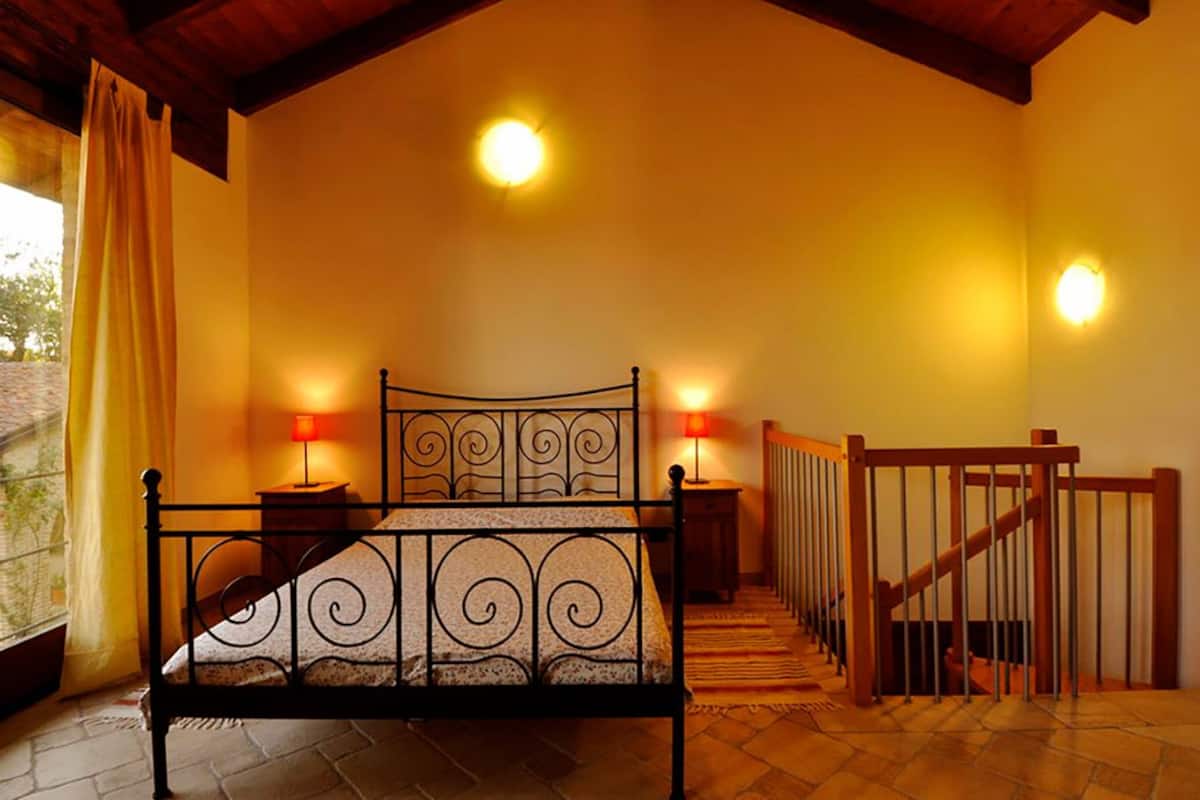 Ein Schlafzimmer mit einem Bett, zwei Nachttischlampen, einer Holztreppe und Blick durch ein Fenster.