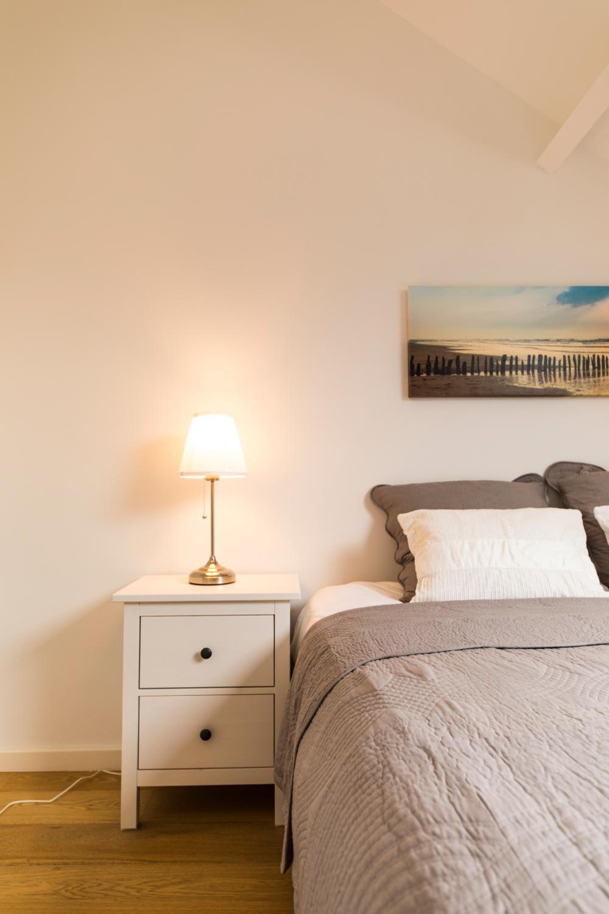 A bedroom with a bed, a bedside table with a lamp, a wall-mounted painting, and a white nightstand with drawers.