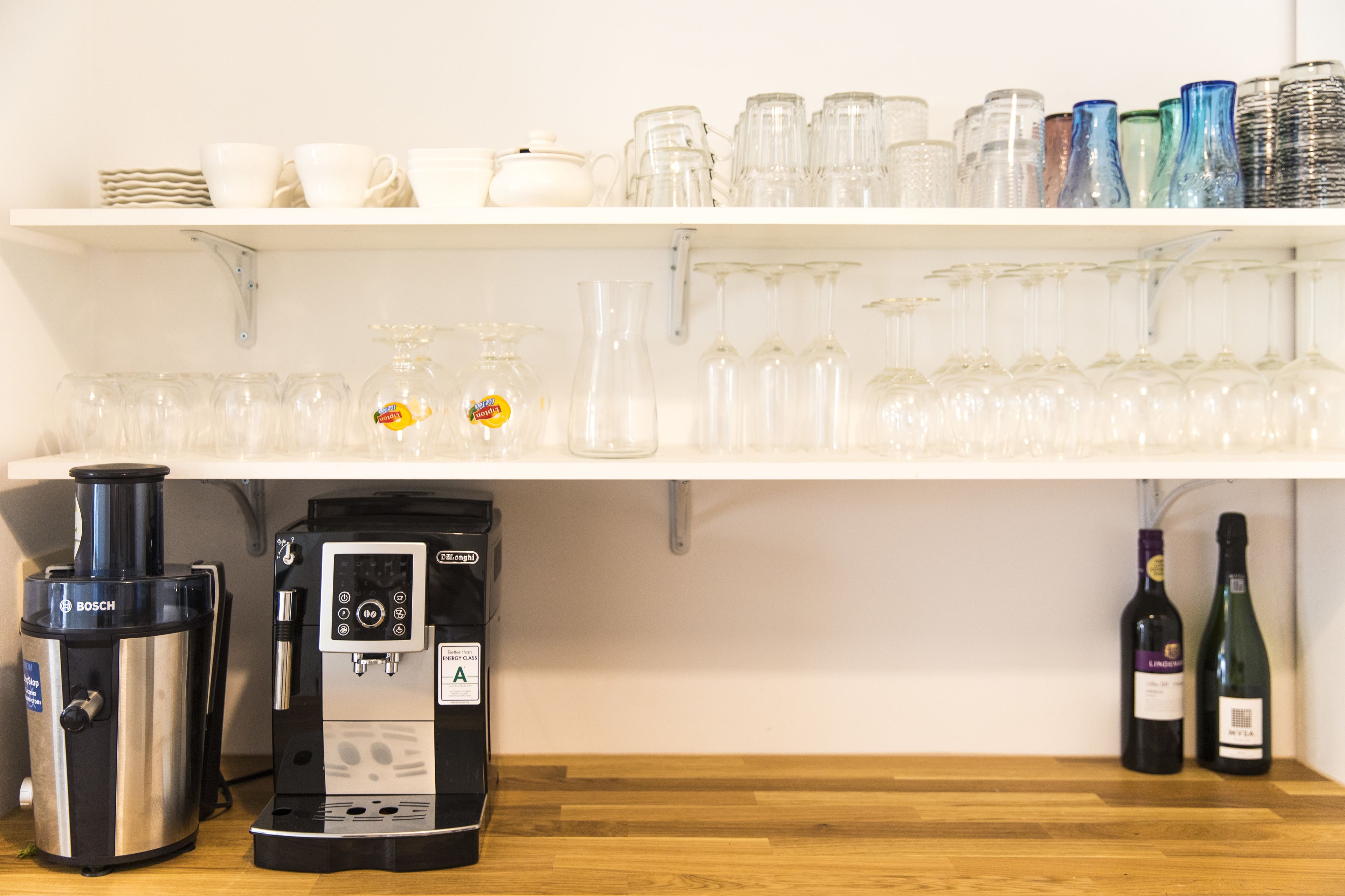 Een keukenhoek met een koffieapparaat, wijnflessen en diverse glazen op planken.