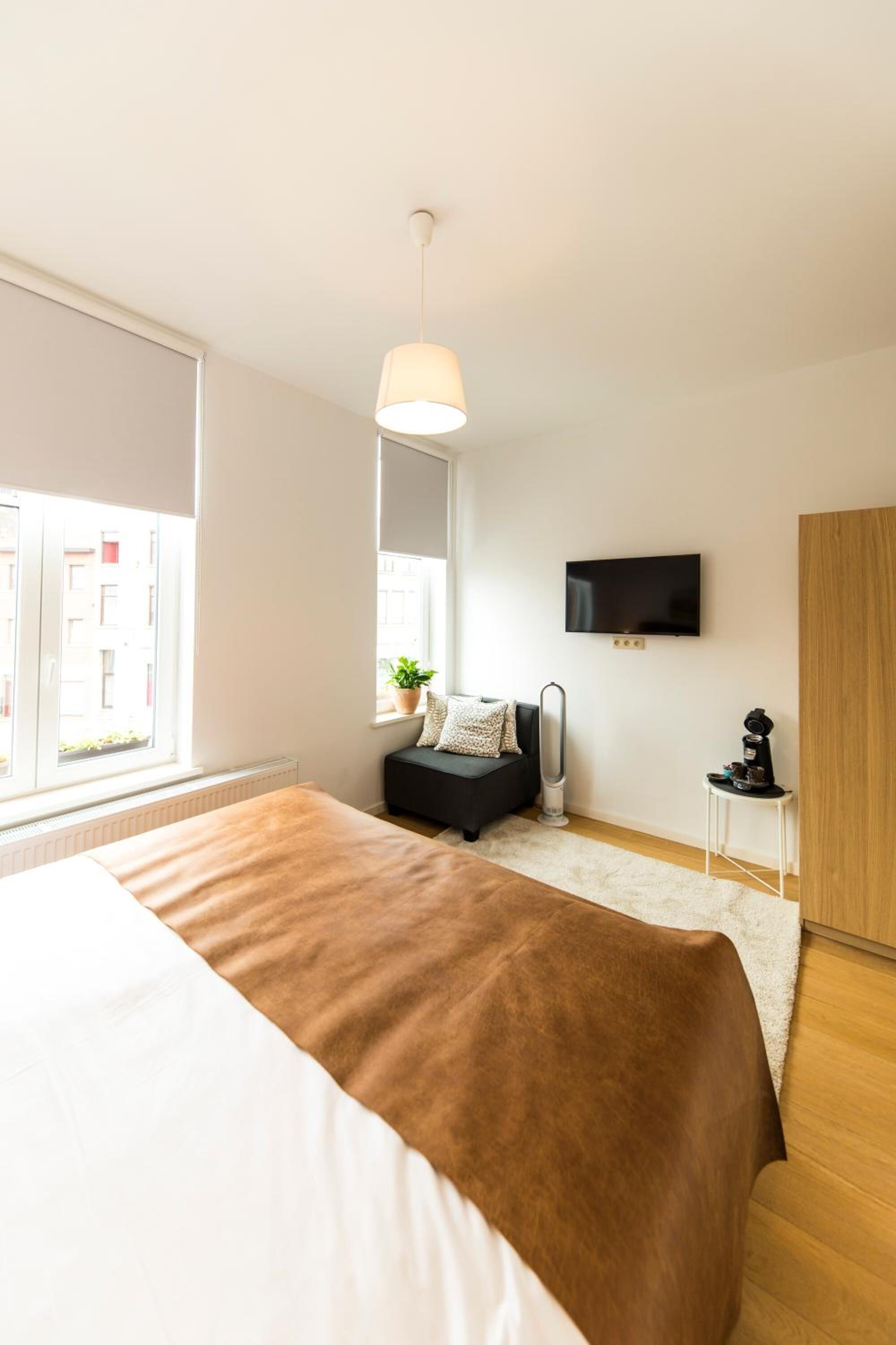 A modern bedroom with a large bed, a TV mounted on the wall, a small table with a plant, and a radiator.