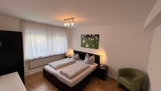 A bedroom with a bed, two bedside tables, a nightstand, a green armchair, a window with sheer curtains, and a wall-mounted light fixture. - Apartmenthaus Somborn (Bochum)