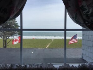 Vista pela janela com duas bandeiras, canadense e americana, sobre uma praia com um caminho gramado.