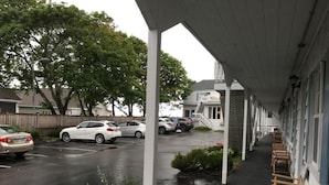A covered outdoor area with a row of wooden chairs, a parking lot with cars, and a building with a white facade in the background. - Holiday House Inn (Scarborough)