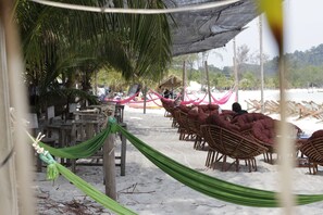 A beachside seating area with wicker chairs and a hammock under a shaded area, with a thatched hut in the background. - Sandbank Restaurant & Camping Koh Rong (Koh Rong)