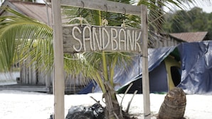 A wooden sign with the word "SANDBANK" in capital letters, a palm tree, and a tent in the background. - Sandbank Restaurant & Camping Koh Rong (Koh Rong)