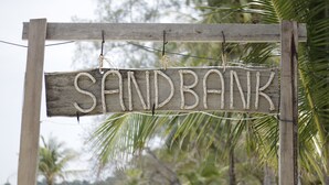 A wooden sign with the word "SANDBANK" written in a rustic font, hanging on a wooden frame with ropes, set against a backdrop of palm trees. - Sandbank Restaurant & Camping Koh Rong (Koh Rong)