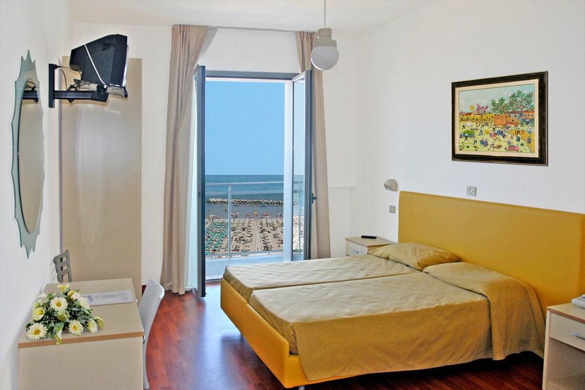 A hotel room with a bed, a desk, a chair, a TV, a painting, and a view of the beach.