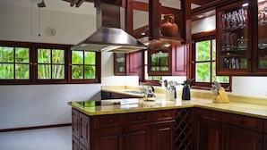 A kitchen with wooden cabinets, a stainless steel range hood, a large window with a view of greenery, and a countertop with kitchen utensils. - Villa Alondra by Casa de Campo Resort & Villas (La Romana)
