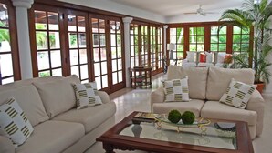 A spacious living room with two sofas, a coffee table, and a view of lush greenery through large windows. - Villa Alondra by Casa de Campo Resort & Villas (La Romana)