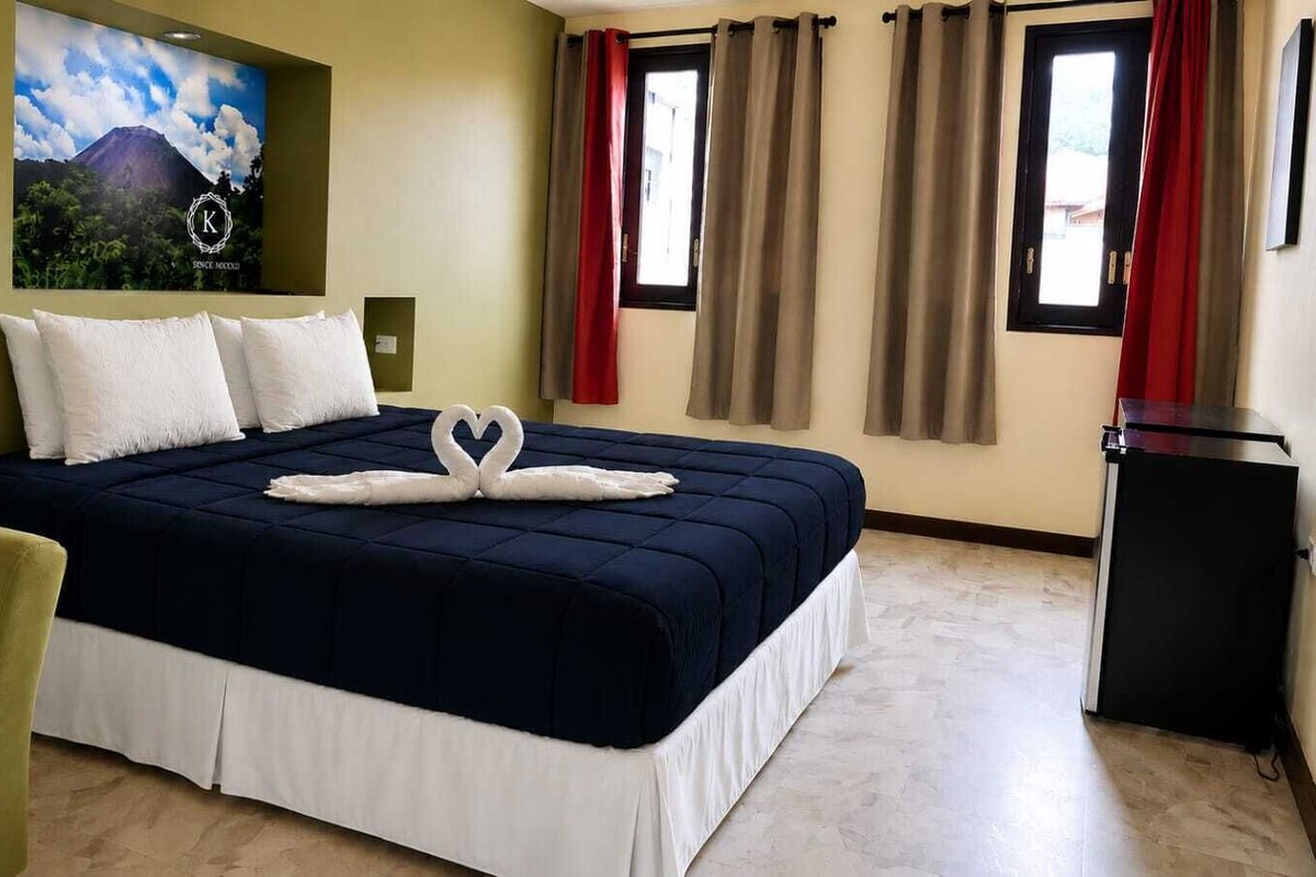 Ein Hotelzimmer mit einem Bett, einem Schreibtisch, einem Sessel, einem Kühlschrank und einem an der Wand befestigten Fernseher.