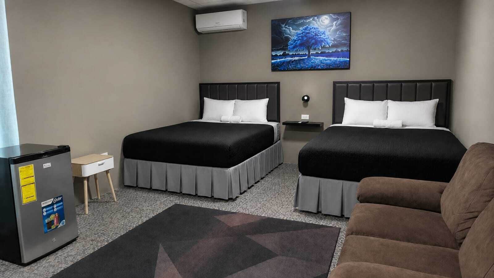 Quarto de hotel com duas camas, um refrigerador, uma mesa pequena, um sofá e um quadro na parede.