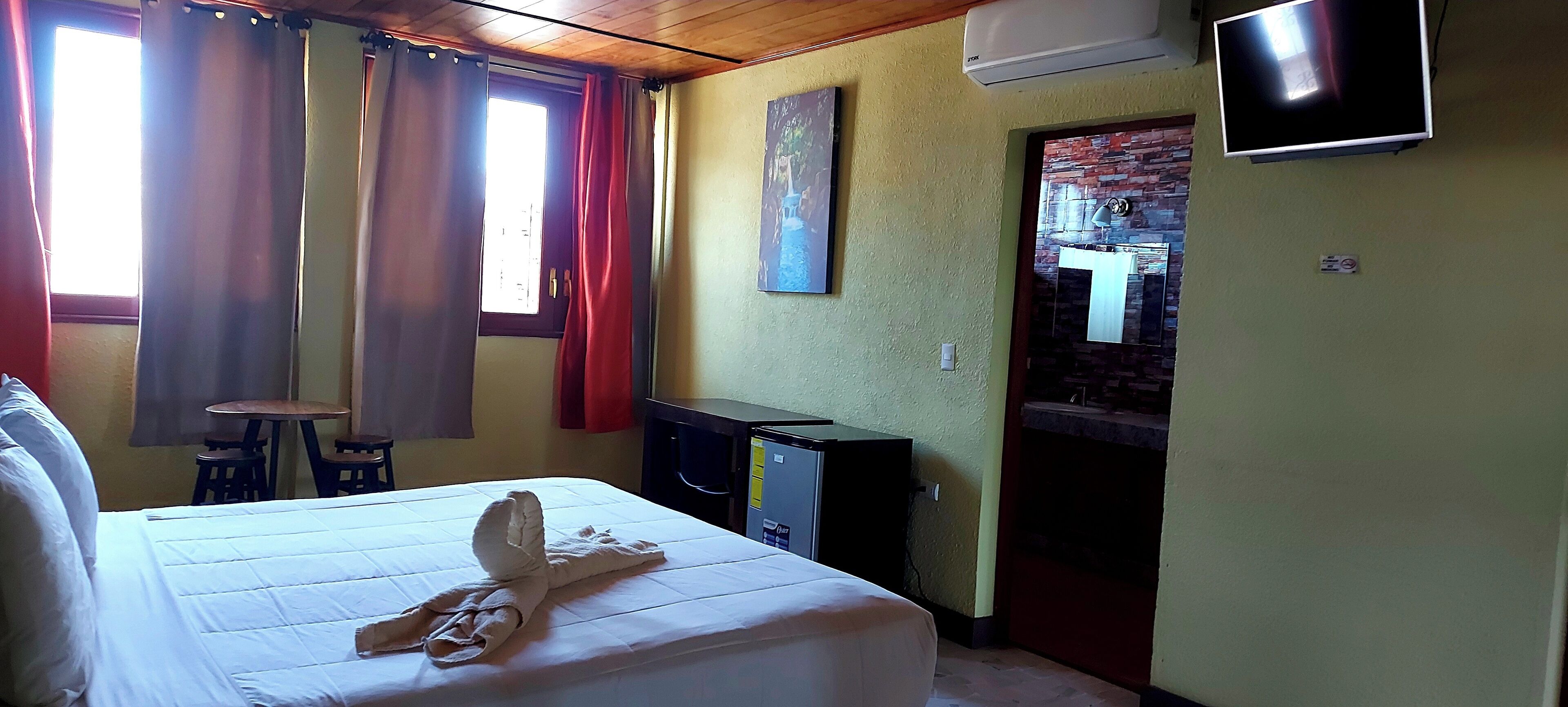 A hotel room with a bed, a desk, a chair, an air conditioning unit, a television, and a small table.