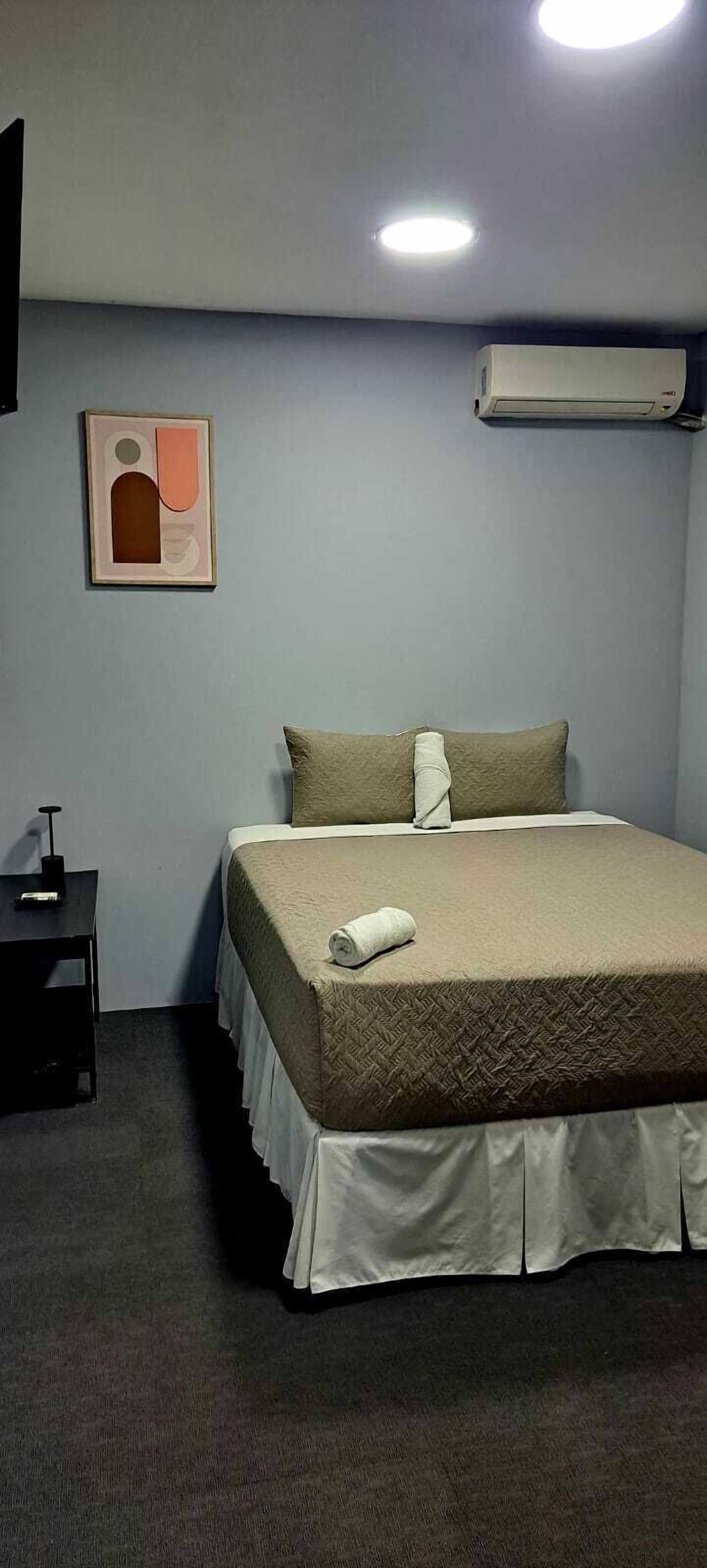 A hotel room with a bed, a pillow, a bedside table, and an air conditioning unit on the wall.