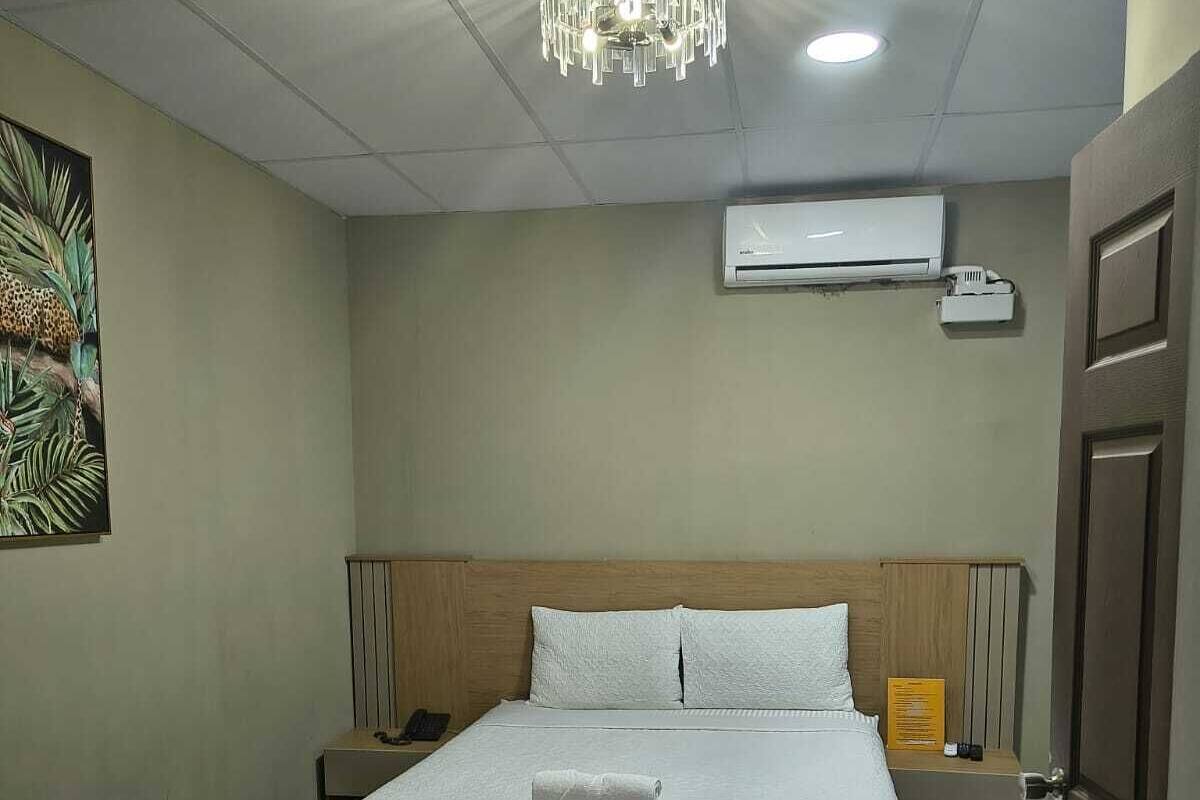 Habitación de hotel con cama, lámpara de araña, unidad de aire acondicionado, mesita de noche y un cuadro en la pared.