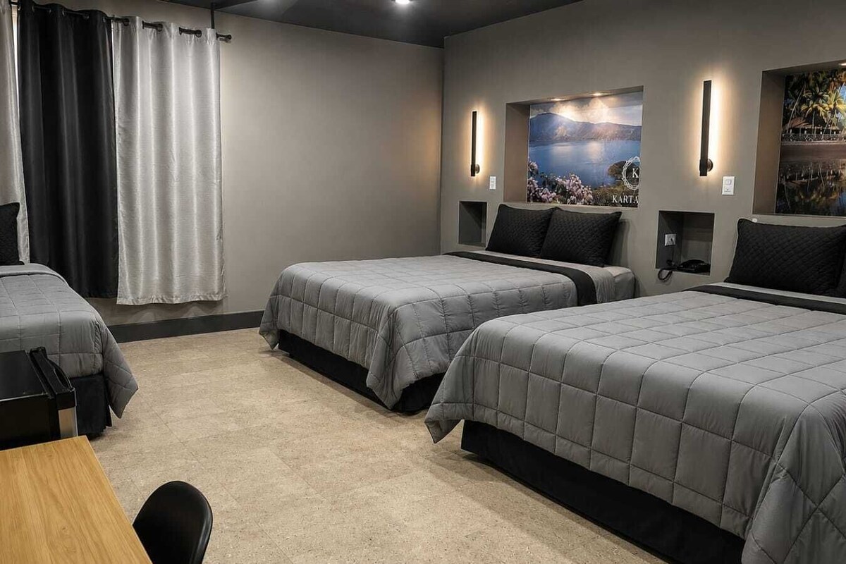 Ein Hotelzimmer mit drei Betten, einem Holztisch, einem Stuhl und einem großen Fenster mit Vorhängen.