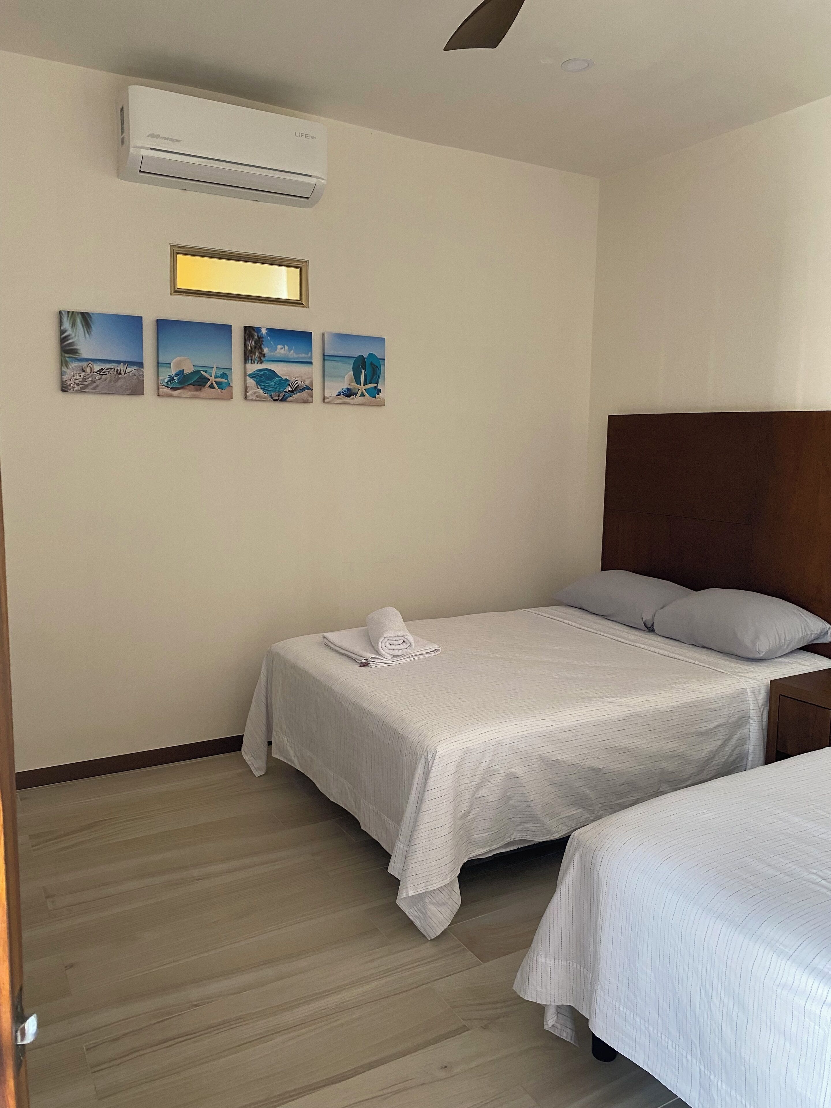 Una habitación de hotel con dos camas, un cabecero de madera, un aire acondicionado de pared y varias pinturas con motivos playeros.