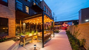 An outdoor patio area with seating and string lights, adjacent to a building with a brick facade and a balcony.