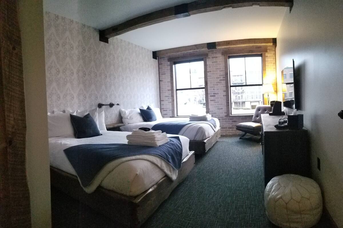 Quarto de hotel com duas camas, uma escrivaninha, uma cadeira, uma luminária e uma janela grande com vista para a cidade.